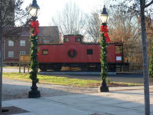 A red caboose decorated with a wreath, surrounded by festive greenery and lampposts, set in a park-like area.