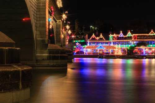 Colorful holiday lights reflect on water at night, illuminating festive buildings along the shore.