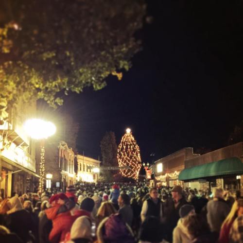 A festive street scene at night, bustling with people and illuminated by a large Christmas tree and holiday lights.