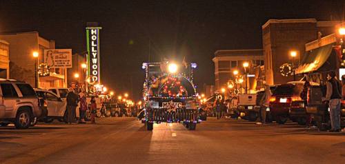 A festive parade scene at night, featuring a decorated vehicle and illuminated storefronts along a bustling street.