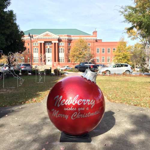 A large red Christmas ornament with "Newberry wishes you a Merry Christmas!" in front of a historic building.