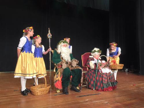 A festive stage scene with children in traditional costumes and a bearded man in green, seated with a woman in a floral crown.