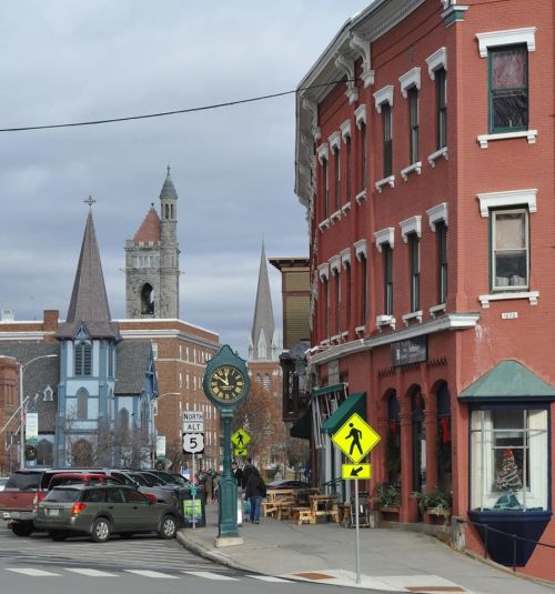 A quaint street scene featuring historic buildings, a clock tower, and cars parked along the road under a cloudy sky.