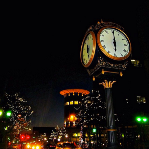 A decorative clock stands in a city at night, surrounded by twinkling lights and a modern building in the background.