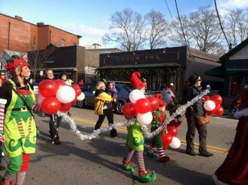 A festive parade scene with participants in colorful costumes holding balloons and decorations, walking down a street.