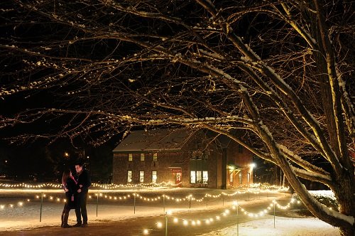A couple stands together under a tree, surrounded by glowing lights and a snow-covered landscape at night.