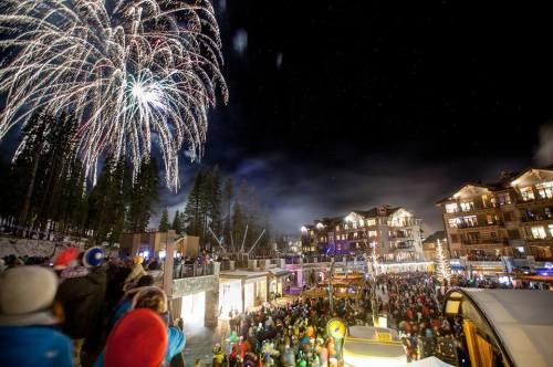 Fireworks illuminate the night sky over a festive crowd in a snowy mountain village, with buildings and trees in the background.
