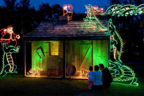 A festive scene with colorful holiday lights on a cabin, featuring Santa and alligators, as two children admire the display.