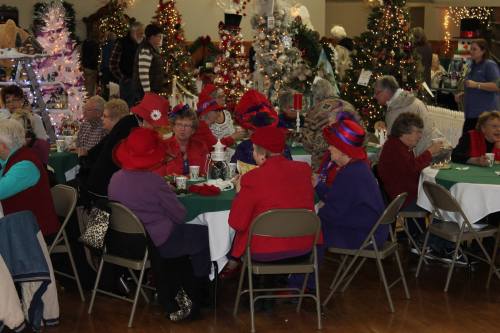 A group of elderly women in colorful hats gather around tables decorated for a festive event, surrounded by Christmas trees.