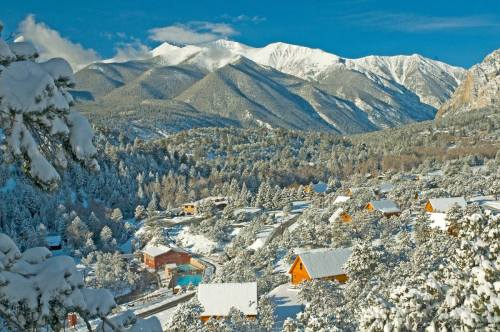 Snow-covered mountains rise above a picturesque valley dotted with cozy cabins and evergreen trees.