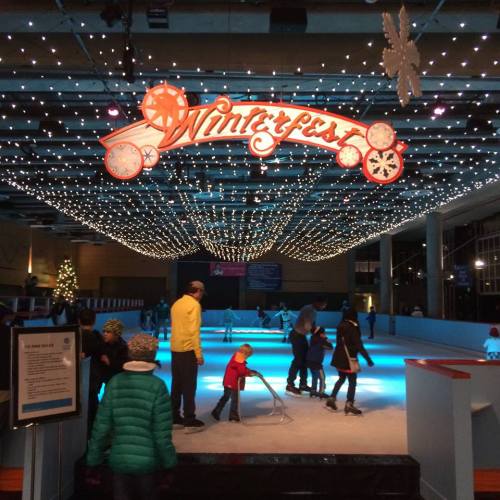 A festive ice skating rink decorated with lights and a "Winterfest" sign, with people skating and enjoying the atmosphere.
