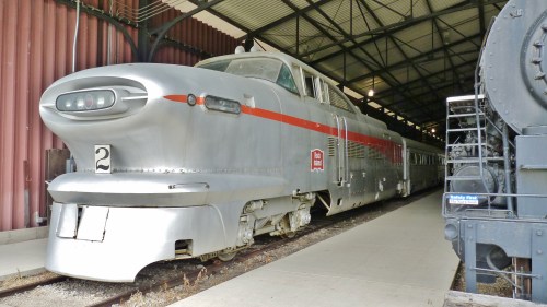 A vintage silver train with a streamlined design, parked inside a train shed next to a black steam locomotive.