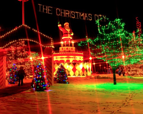 Colorful holiday lights illuminate a festive scene with a large Santa figure and decorated trees in a snowy setting.