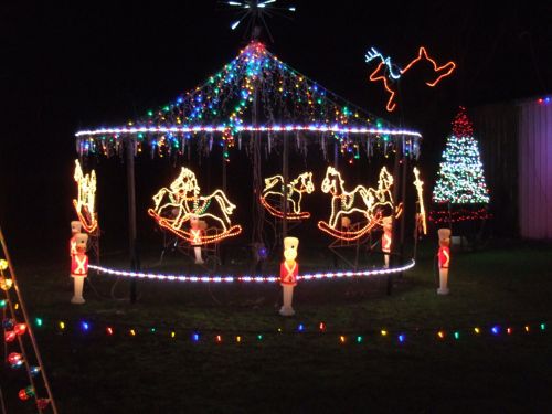 A festive display featuring a carousel with illuminated horses, surrounded by colorful lights and a decorated Christmas tree.