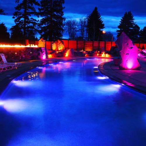 A serene pool at night, illuminated with colorful lights and surrounded by trees and stone features.