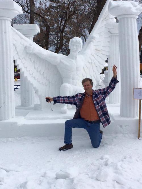 A man kneels beside a large snow sculpture of a winged figure with columns in a snowy park setting.