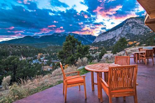 A scenic view of mountains and colorful clouds at sunset, with wooden chairs and a table on a patio.
