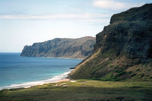 A scenic coastal view featuring rugged cliffs, sandy beaches, and calm blue waters under a partly cloudy sky.