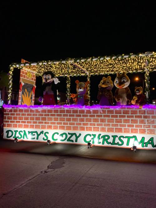 A festive parade float decorated with lights, featuring Disney characters in a cozy Christmas theme at night.