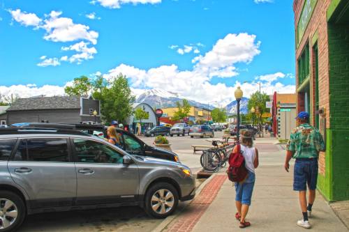A sunny street scene with people walking, shops, parked cars, and mountains in the background under a blue sky.