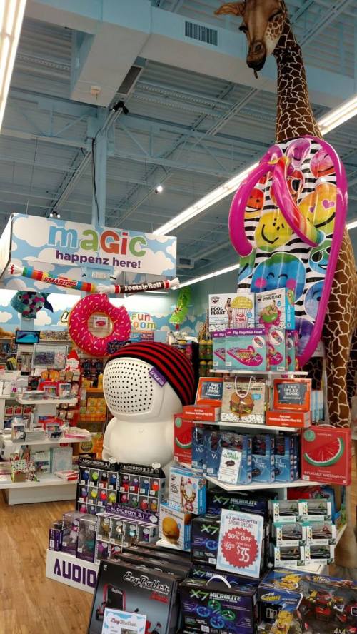 A colorful store display featuring toys, games, and a large giraffe statue, with a sign reading "Magic happens here!"