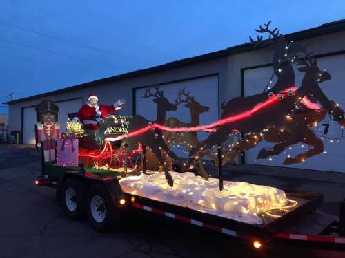 A festive float featuring Santa Claus, reindeer, and holiday lights, set against a twilight sky.