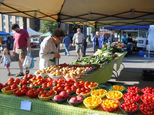 A farmer's market stall displays colorful fruits and vegetables, with a woman browsing the produce.