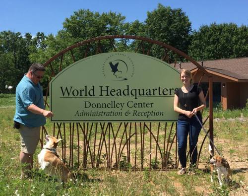 Two people stand beside a sign that reads "World Headquarters" with two dogs on leashes in a grassy area.