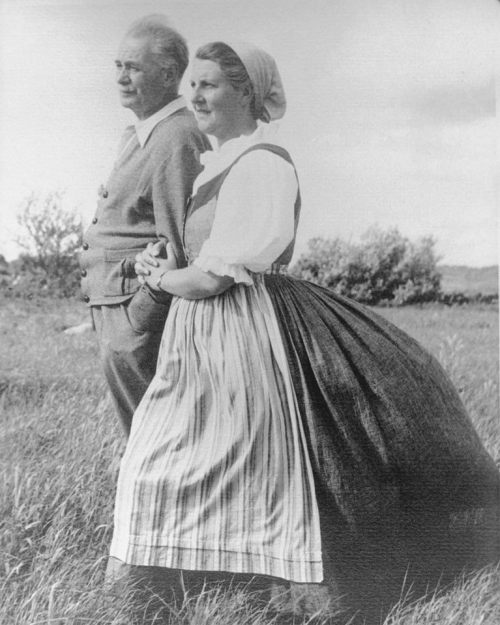 A man and woman stand together in a grassy field, dressed in vintage clothing, with a serene landscape in the background.