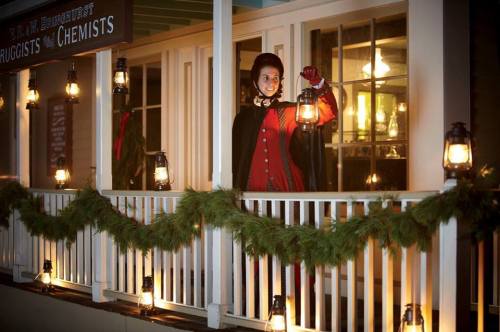 A mannequin in a red cape holds a lantern on a porch decorated with greenery and lanterns, evoking a festive atmosphere.