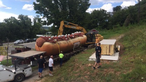 A large hot dog sculpture being loaded onto a flatbed truck with a yellow excavator, surrounded by people and trees.