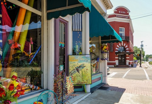 Colorful storefront with decorations and artwork, featuring a bank building in the background on a sunny day.