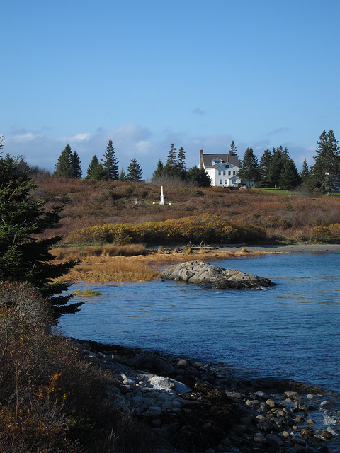 A serene coastal scene featuring a white house on a hill, surrounded by trees and golden grasses, with a calm blue water foreground.