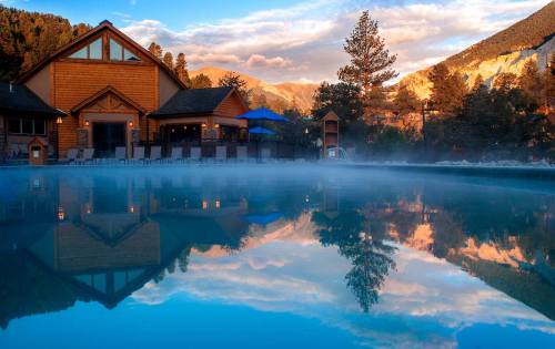 A serene pool reflects a mountain lodge and colorful sky, surrounded by mist and trees.