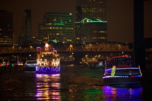 Colorful boats adorned with lights navigate a river at night, with a city skyline and bridge in the background.