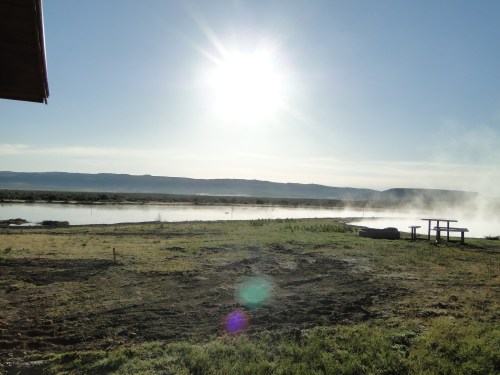 A serene landscape featuring a river, mountains in the background, and steam rising in the sunlight.