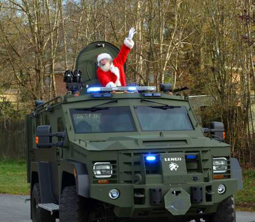 Santa Claus waves from the top of a military-style vehicle, adorned with lights, in a wooded area.