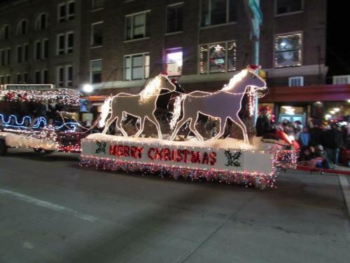 A festive parade float featuring two illuminated horses and the message "Merry Christmas," surrounded by colorful lights.