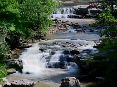A serene river flows over rocks, surrounded by lush green trees and gentle waterfalls in a peaceful natural setting.