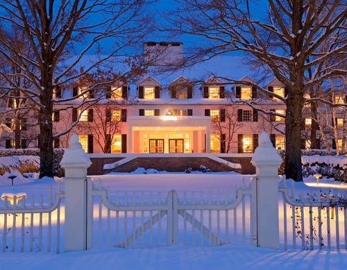 A grand house illuminated at dusk, surrounded by snow and trees, with a white fence in the foreground.