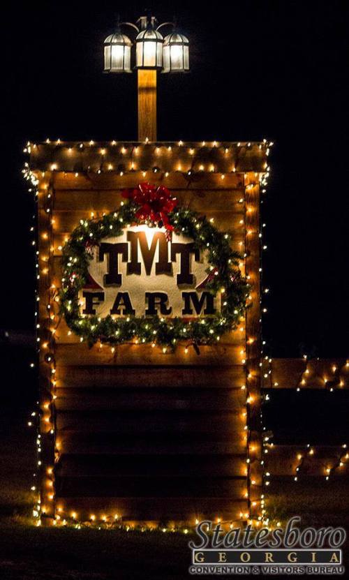 A wooden sign for TMT Farm, decorated with a wreath and string lights, illuminated at night.