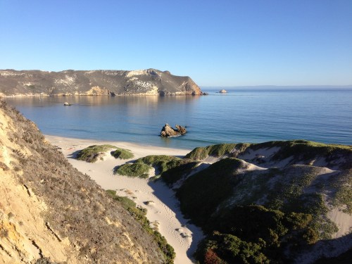 A serene beach scene with gentle waves, rocky formations, and lush green hills under a clear blue sky.