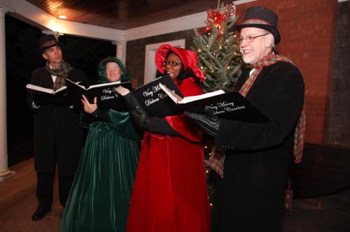 Four carolers in festive attire sing together, holding songbooks, with a decorated Christmas tree in the background.