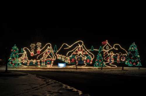 Colorful holiday lights outline a whimsical village scene with trees, mountains, and festive decorations against a night sky.