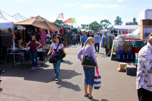 A bustling outdoor market with vendors, shoppers, and colorful tents under a clear blue sky.