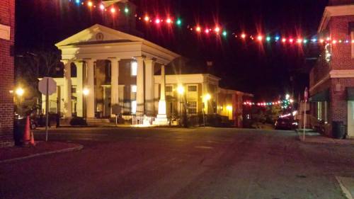 A quiet street at night, featuring a lit building with columns and colorful string lights overhead.