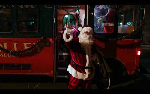 A cheerful Santa Claus waves from the doorway of a decorated trolley bus, spreading holiday cheer.