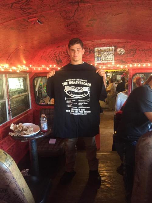 A young man holds a black t-shirt with a graphic design inside a decorated bus. Warm lights illuminate the space.