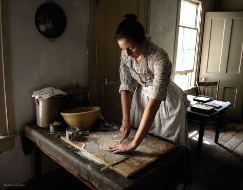 A woman in period clothing kneads dough on a wooden table in a rustic kitchen with natural light.