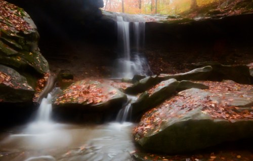A serene waterfall cascades over rocks, surrounded by autumn leaves in a tranquil forest setting.
